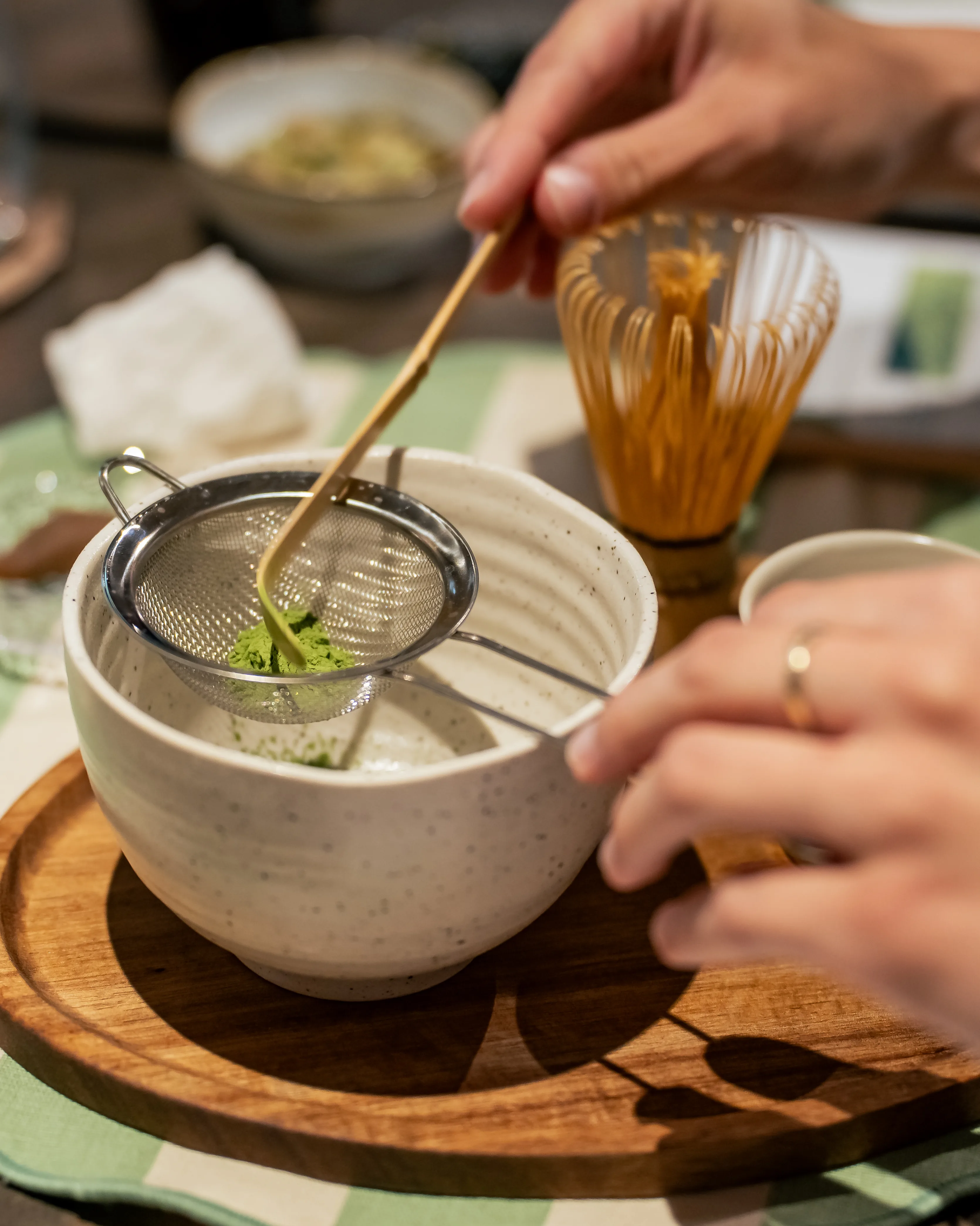 Water poured from a dark green ceramic pourer into a small matcha cup, the liquid catching the light over dried tea leaves and the natsume container