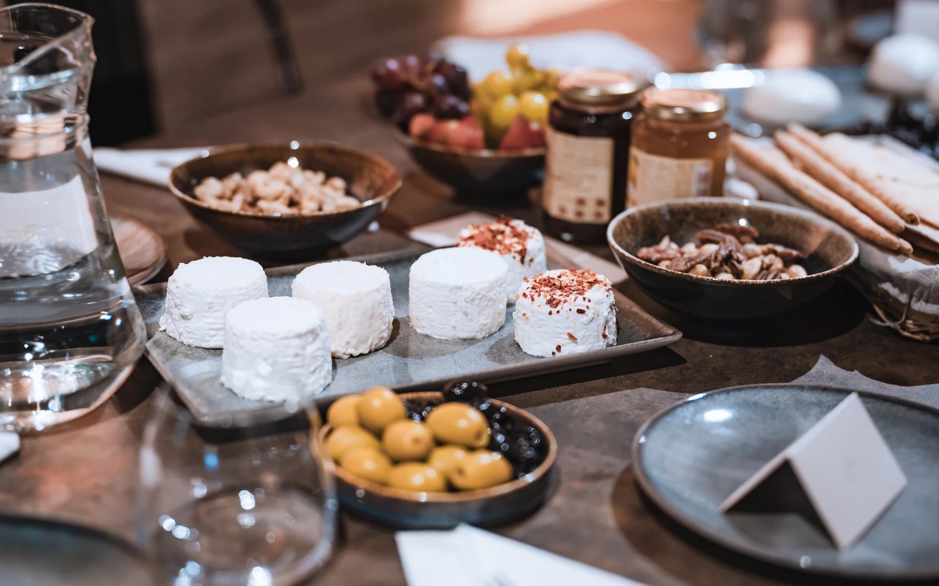 Fresh white cheeses on a stone tray, two topped with chilli flakes, among bowls of olives, nuts and grapes and jars of preserves
