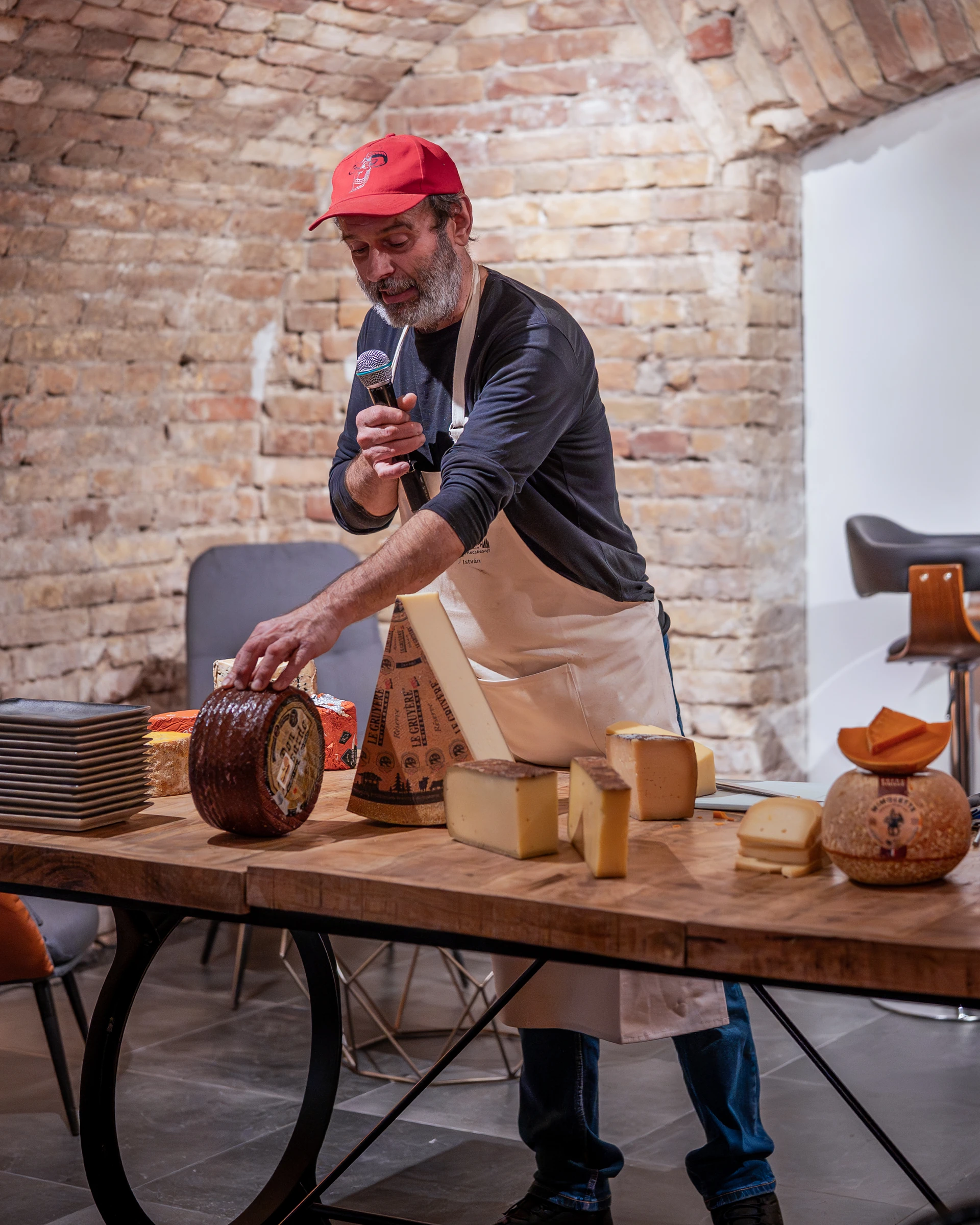 István Csirmaz, microphone in hand, resting a hand on a waxed cheese wheel beside a wedge of Gruyère and other hard cheeses under the brick vault