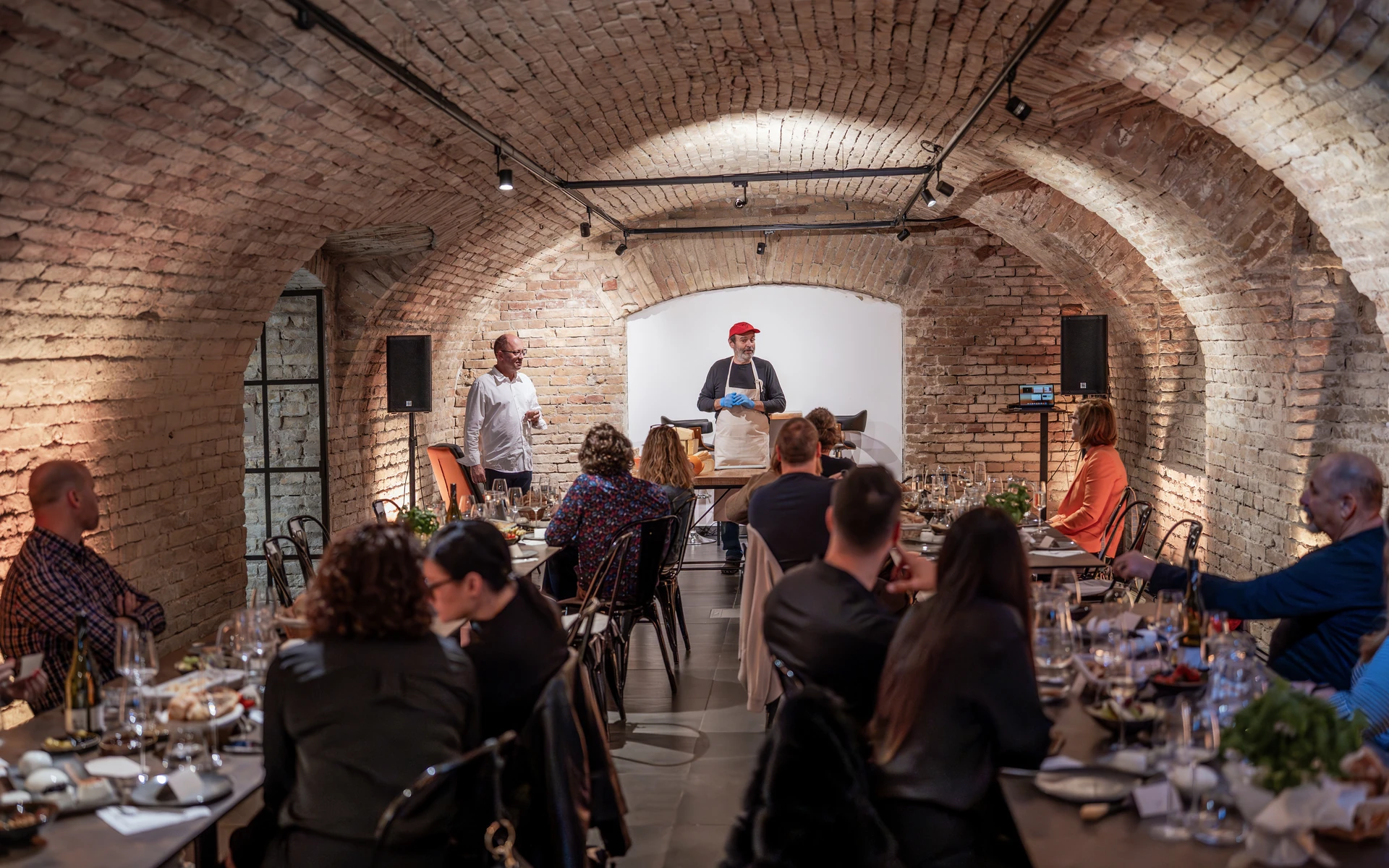 Forty-one guests at two long tables beneath the vaulted brick ceiling, facing the cheese master at the far end of the cellar